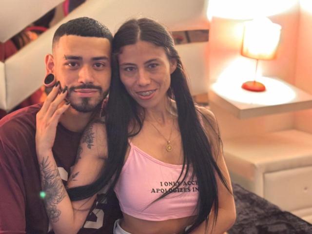 TashaAndRolo with long black hair, pink crop top, small tits, and Rolo with short black hair, beard, tattoos, and ear gauges, holding up a finger, smiling.