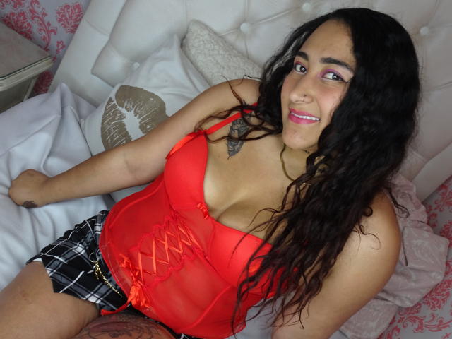 AngelineThomsom with long black hair, red lingerie, plaid shorts, and pink eyeshadow, sitting on a bed, smiling, showing her tits and a small tattoo on her chest.