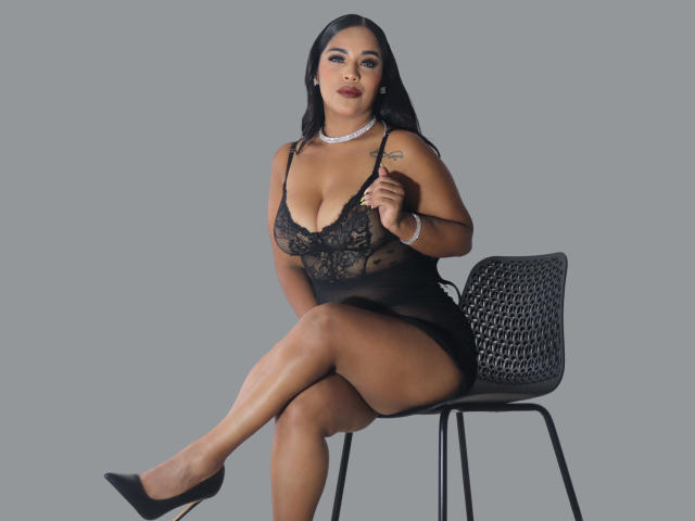 Lana Rossel in schwarzer Spitzenunterwäsche, die ihre großen Brüste zeigt, sitzt auf einem Stuhl, die Beine gekreuzt, trägt schwarze High Heels, eine silberne Halskette und ein Armband, vor einem schlichten grauen Hintergrund..