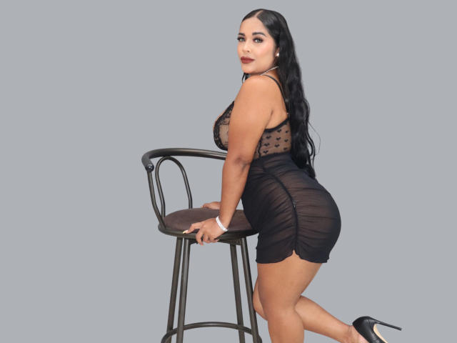LanaRossel with black polka dot sheer top, showing her tits, and tight black skirt, leaning on a stool, black heels, long black hair, looking back, gray background.