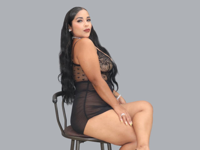 LanaRossel sitting on a chair, wearing a sheer black lace dress, showing her ass, long black hair, looking back, gray background, nude nails, earrings, and a bracelet.