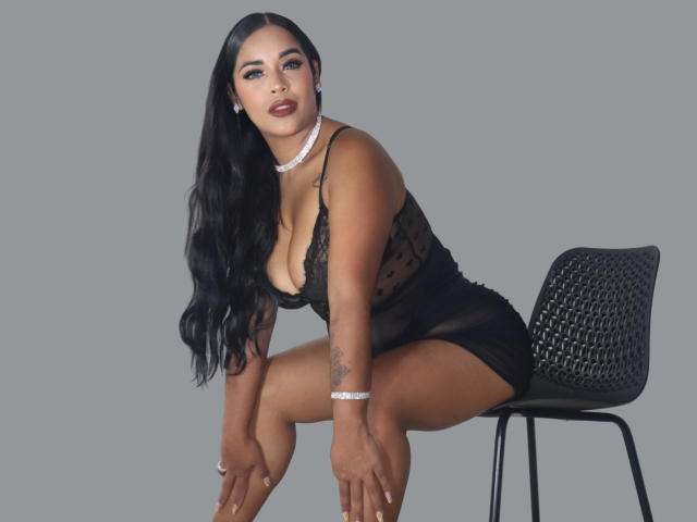 LanaRossel, with black lace dress, sitting on black chair, showing her tits, wearing white choker, long black hair, tan skin, tattoo on right arm, gray background.