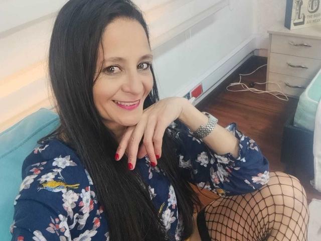 Meredith Hunnt, con cabello negro largo, sonriendo, vestida con una camisa azul con estampado floral, medias de red, esmalte de uñas rojo y un reloj de plata, apoyando su barbilla en su mano..