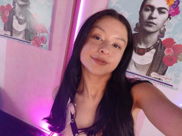DyanaMoon with long black hair, brown eyes, and a slight smile, wearing a purple top, in front of two black-and-white portraits, pink wall, and purple light.