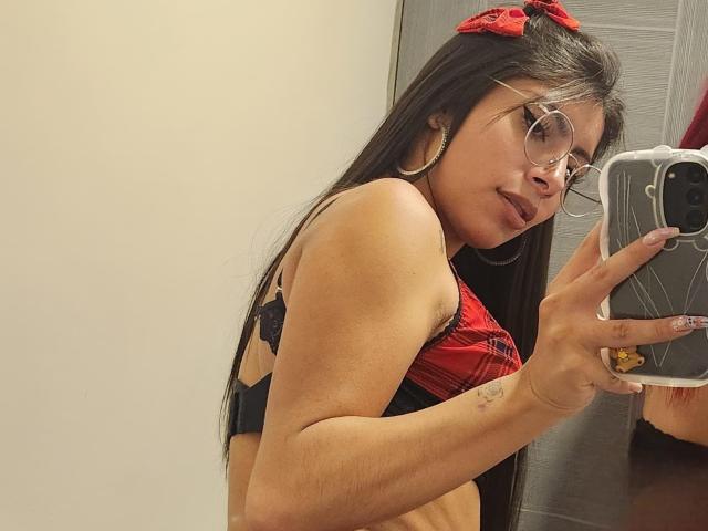 LeiaLaurent with long black hair, red bow, glasses, red plaid bra with black lace, taking a mirror selfie, showing her small tits, holding a phone with a yellow sticker.