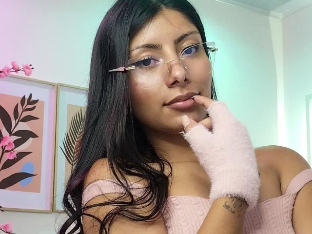 LeiaLaurent ha lunghi capelli neri, occhiali, una maglietta rosa a spalle scoperte e un guanto rosa peloso senza dita, con un dito che tocca le sue labbra. Ha la pelle abbronzata e un tatuaggio..