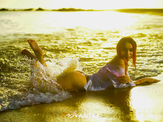 AmyKenny liegt am Strand, trägt ein nasses, weißes Hemd, hat rotes Haar, den Hintern hoch, die Beine im Wasser, die Zehen sind nach vorne gerichtet, Sonnenschein, Wellen, goldene Stunde, Wasser spritzt, sexy, süß, entspannt..