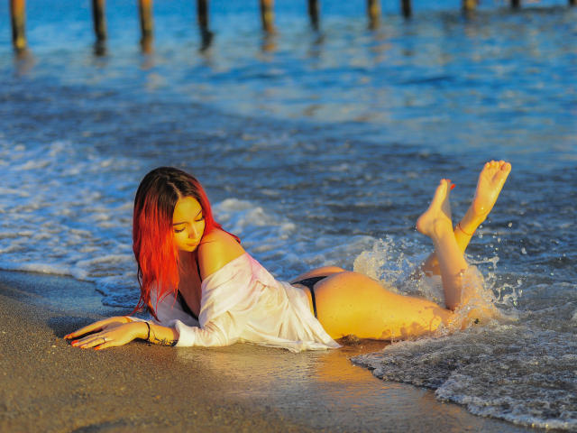 Amy Kenny, con cabello rojo, acostada en una playa, vistiendo una camisa blanca y una tanga negra, con las nalgas levantadas, los pies en el agua, el sol brillando en su piel, con las piernas extendidas hacia atrás..