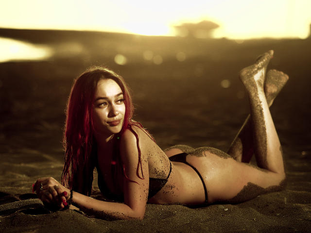 AmyKenny lying on a sandy beach, wearing a black bikini, her red hair, sand-covered legs, and a playful smile. Sunlight highlights her curvy ass and small tits.