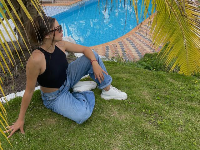 SashaHarding, with small tits, sits on grass by a pool, wearing a black crop top, blue jeans, and white sneakers, looking sideways with glasses, palm leaves in the foreground.