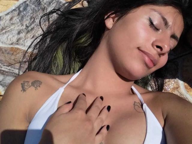 LeiaLaurent with black nails, eyes closed, smiling, sunlit, wearing white bikini, hand on chest, tattoos on shoulder and chest, black hair, tan skin, sunlight on face.