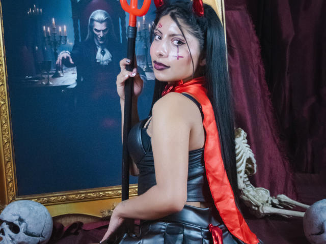 LeiaLaurent com uma blusa de couro preta, lenço vermelho, chifres de diabo e maquiagem, segurando um tridente vermelho, olhando para trás, com uma pintura escura e assustadora e caveiras ao fundo..