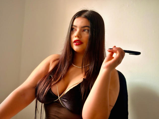 LaraJames with long dark hair, red lipstick, black sheer dress, holding a makeup brush, pearl necklace, large tits, light skin, confident smile, soft light, indoor setting.