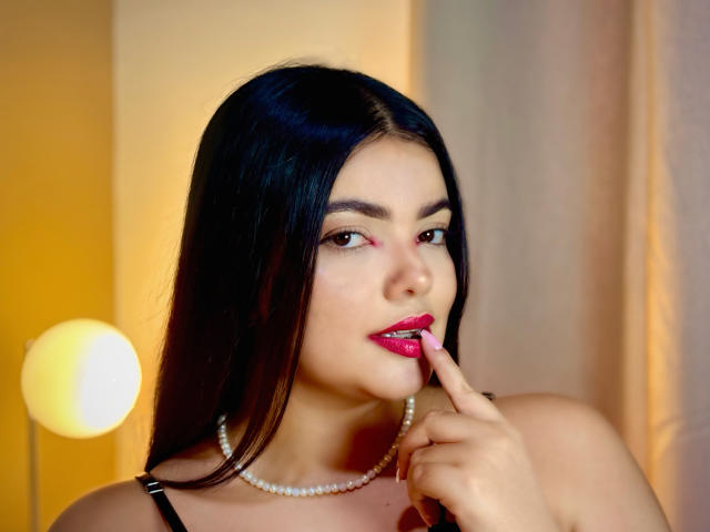 LaraJames with black hair, pink lipstick, and pearl necklace, biting her finger, showing red lipstick on her finger, wearing a black strap top, with a sexy, confident look.