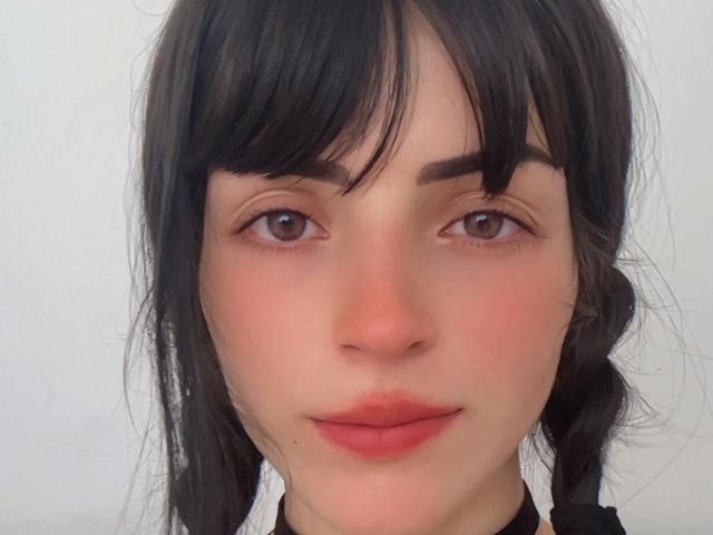 Liiil has pale skin, dark brown eyes, and black, slightly messy hair with bangs and a braid. She's wearing pink lipstick with a subtle smile.