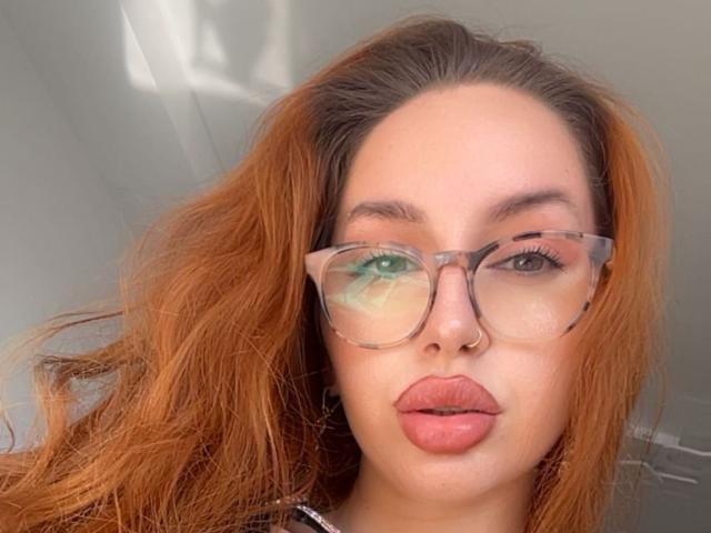 GinnyJoy with reddish-brown hair, full lips, nose ring, and clear glasses, looking directly into the camera with a serious expression.