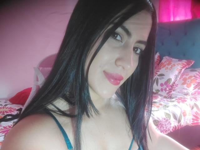 SofiaFountaine with black hair, pink lipstick, and blue bra, smiling at the camera. Her tits are visible, and she has a cute face. Bright pink light in the background.