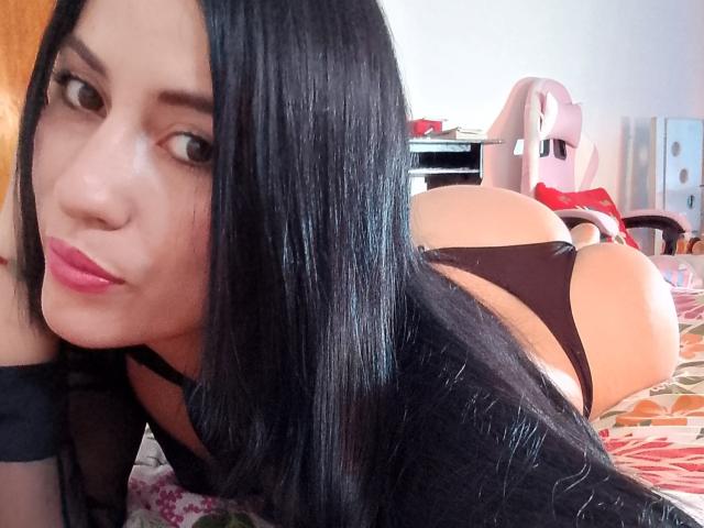 SofiaFountaine with long black hair, pink lipstick, black lingerie, and black sheer sleeves, lying on a bed, butt in the air, looking at the camera.
