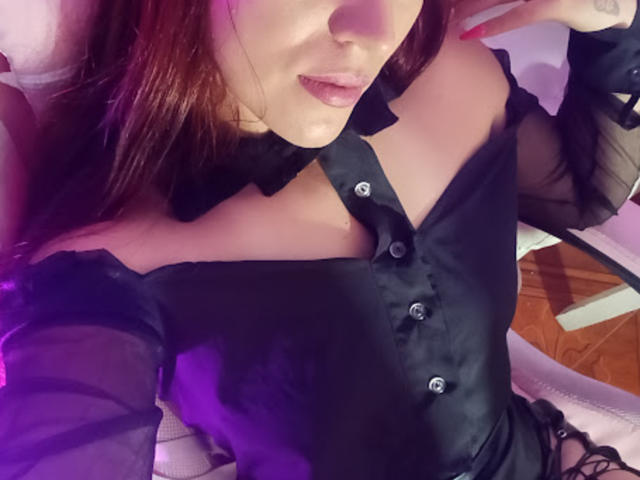 SofiaFountaine with dark hair, wearing a black, off-shoulder, button-up dress with sheer sleeves, showing her cleavage, and a hint of black lace underwear, in a purple-lit room.
