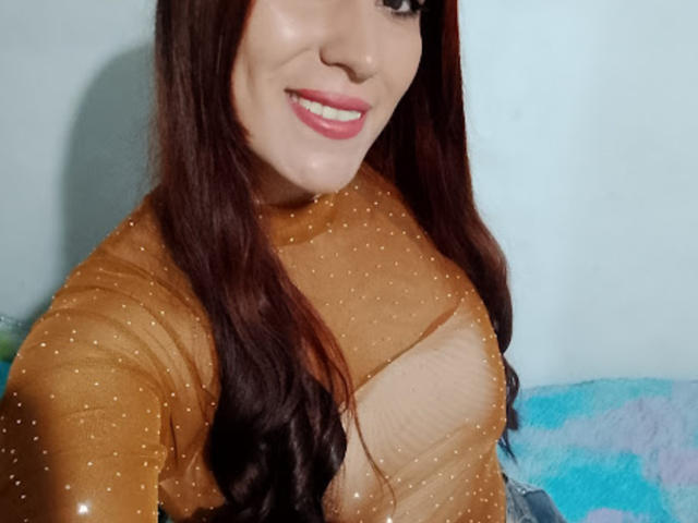 SofiaFountaine with red hair, wearing a sheer, brown, polka-dotted top, showing her tits through the fabric, pink lips, and a light blue wall in the background.
