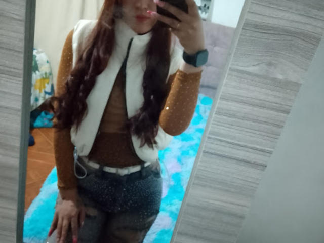 SofiaFountaine with long brown hair, smiling, wearing a sheer brown top that shows her small tits and nipples. She's got pink lipstick and is looking at the camera.