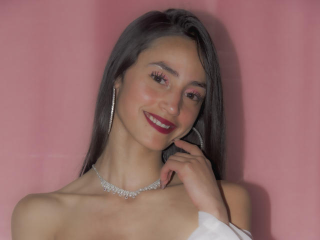 Meganoa with medium tits, wearing a white off-shoulder top, diamond necklace, and hoop earrings, smiling with red lipstick, holding her right hand, against a pink background.