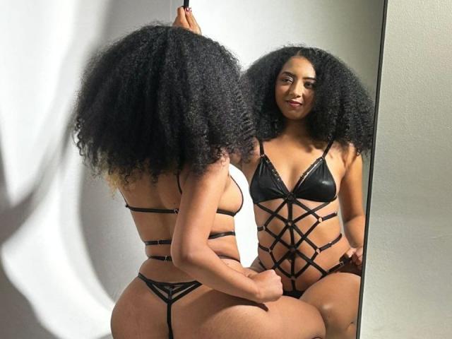 NahomiMeilleur with curly black hair, wearing strappy black lingerie, sits in front of a mirror. She has a medium brown skin tone, small boobs, and a curvy ass.