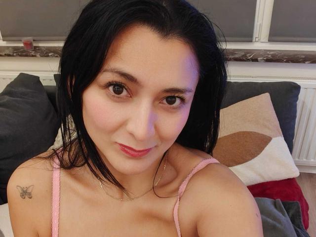 CharlotteGossart with black hair, pink spaghetti strap top, butterfly tattoo on right shoulder, pink lipstick, subtle smile, medium tits, light skin, brown eyes, indoor, selfie.