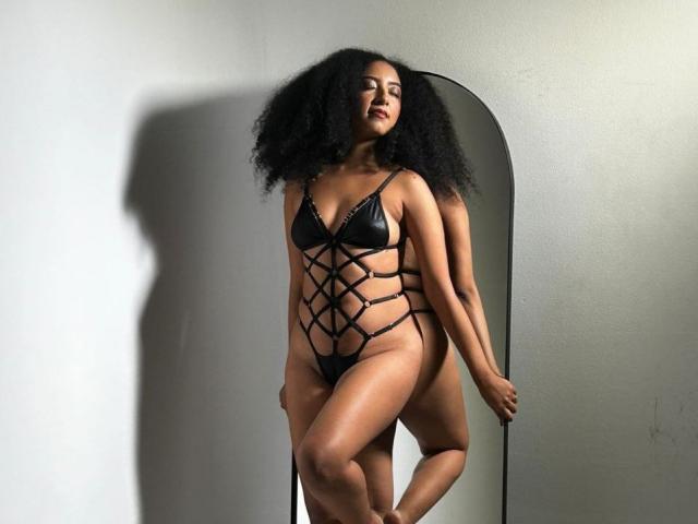 NahomiMeilleur, con i capelli neri ricci, indossa un set di lingerie nera con spalline sottili, che mette in mostra i suoi seni e i suoi genitali, ed è in piedi accanto a uno specchio, mostrando il suo riflesso..