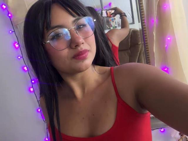 NovaAllen mit schwarzen Haaren, blauen, spiegelnden Brillen und einem roten Oberteil, macht ein Selfie und zeigt ihre Brüste, ihre rosafarbenen Lippen und im Hintergrund lila Lichterketten..