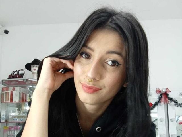 AlisKooper with long black hair, nose ring, and pink lipstick, smiling slightly, touching her hair with her right hand, wearing a black shirt.