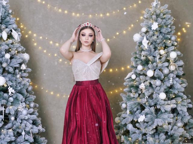 AmandaGrace stands between two snow-dusted Christmas trees, wearing a sparkling white strapless top and a red, pleated skirt. She holds a silver tiara with red jewels in her hands. Her makeup is bold, with dark eyeshadow and pink lipstick. The trees are decorated with white ornaments and lights. AmandaGrace's long brown hair is parted in the middle. The background has a string of warm yellow fairy lights. The image is colorful and festive.