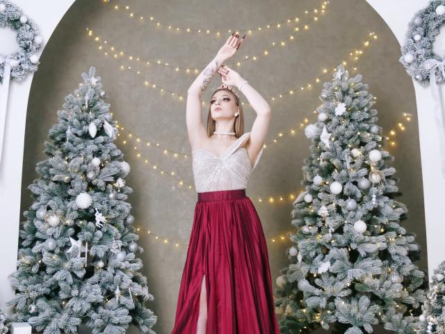 AmandaGrace stands between two frost-covered Christmas trees, arms raised. She wears a white, silver-embellished sleeveless top and a deep red, high-slitted skirt. Her blonde hair is down, and she has a silver headband. The trees are decorated with white and silver ornaments, and warm string lights form a star pattern behind her. The photo is sharp, with a focus on AmandaGrace's elegant pose and festive attire.
