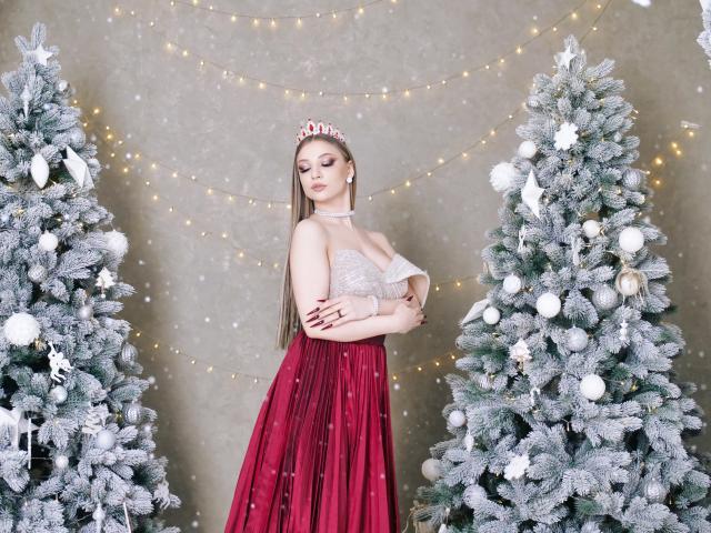 AmandaGrace stands between two frosted Christmas trees adorned with white ornaments and fairy lights. She wears a glittery silver top with a deep neckline and a flowing red skirt. Her long brown hair is straight, and she has a red and white tiara on her head. Her eyes are closed, and she has a serene expression. AmandaGrace's arms are crossed over her chest, and her red nails are visible. The image is bright with a soft, snowy texture and warm fairy lights in the background.