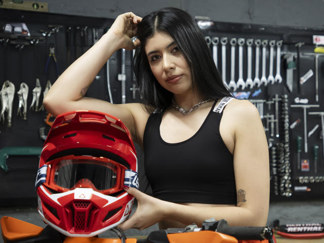MiluSky with black tank top, holding red motorcycle helmet, dark straight hair, subtle tattoo on right arm, wearing chain necklace, in garage workshop, looking at camera.