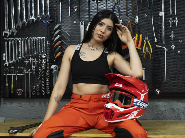 MiluSky con reggiseno sportivo nero, pantaloni rossi e casco rosso, seduta su una panchina, con una mano tra i capelli, che mostra un tatuaggio sul braccio destro e un piccolo tatuaggio sul petto, in una stanza piena di attrezzi..