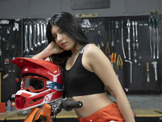 MiluSky with long black hair, wearing a black sports bra and orange pants, leaning on a red motorcycle helmet in a garage. Her tits are small, her ass is not visible.