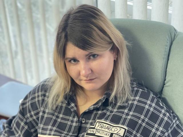MinistraX has blonde and brown hair, wearing a black and white checkered shirt, looking at the camera with a slight frown. She's sitting in a green chair.
