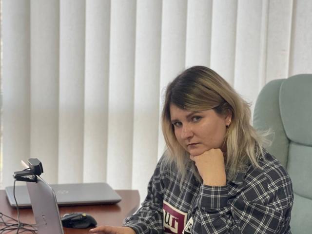 MinistraX, loira com raízes escuras, vestindo uma camisa xadrez, sentada à mesa, com a mão no queixo, com uma expressão séria, um laptop à sua frente, persianas verticais ao fundo, com uma expressão ligeiramente irritada..