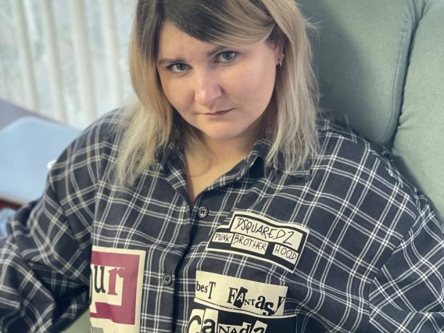 MinistraX with blonde hair, blue eyes, and a serious expression, wearing a black and white plaid shirt, sitting on a green chair, looking straight at the camera.