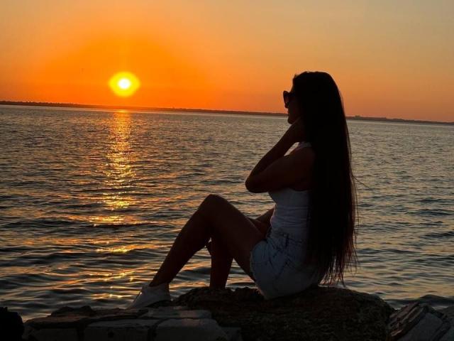 Silhouetted Viollia with long dark hair, white tank top, and denim shorts, sitting on rocks, hand near mouth, sunset over water, orange sky, reflection on water.