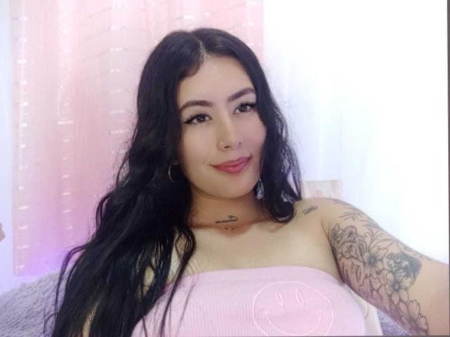 AlisonKweel with long black hair, tattoos on her arm and chest, nose ring, and pink tube top, smiling at the camera, showing her tits.