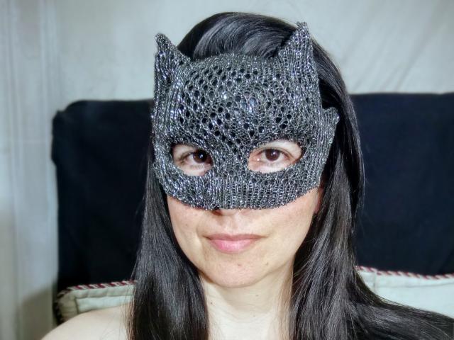 A close-up of RossanaHairy with a detailed black lace mask covering her eyes. The mask has a textured, intricate lace pattern with small, sparkling beads. RossanaHairy has straight, dark brown hair, and a fair skin tone. Her lips are slightly parted, showing a subtle smile. The background is blurred, with a dark, cushioned headrest visible behind her. The lace mask has small, pointed ears at the top.