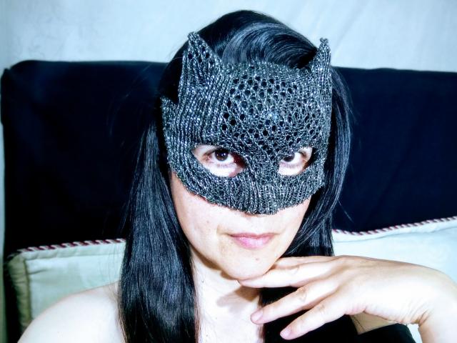 RossanaHairy with a black, net-like mask covering her eyes, has long black hair. She's looking slightly to the side, with her right hand touching her chin. Her skin is fair, and she has a slight, neutral expression. The mask's texture is intricate, resembling a spiderweb. She's in a close-up shot, with a dark background and white pillow behind her. The image has a sharp, high-contrast look.
