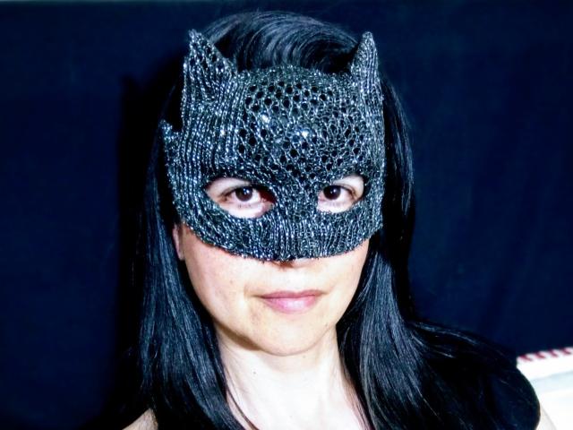 RossanaHairy wears a black, glittery cat mask with pointed ears. Her long, straight black hair frames her face. The mask covers her eyes, leaving only a small part of her brown eyes visible. She has a subtle, closed-mouth smile. The mask's texture is rough and sparkling, contrasting with her smooth, fair skin. The background is dark, making RossanaHairy's mask and hair stand out. The image is clear and focused.