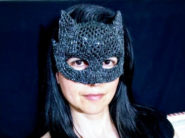 Close-up of RossanaHairy wearing a black, textured, cat-eye mask with intricate bead-like patterns. She has straight, black hair and light skin. Her eyes are visible through the mask, and she's smiling slightly. The mask has a metallic sheen and detailed, raised texture. RossanaHairy's face is partially covered, but her natural expression is visible. The background is dark and indistinct.