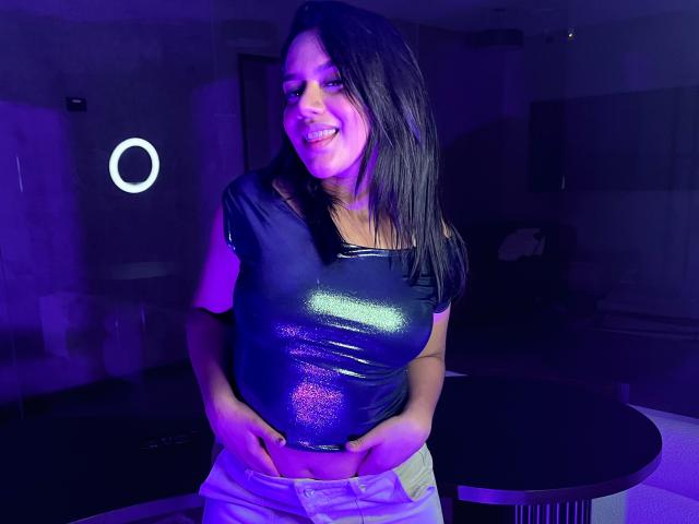 AraFerrer with shiny black top, white pants, hands on her crotch, smiling, purple lighting, black hair, medium tits, standing, ring light in background.
