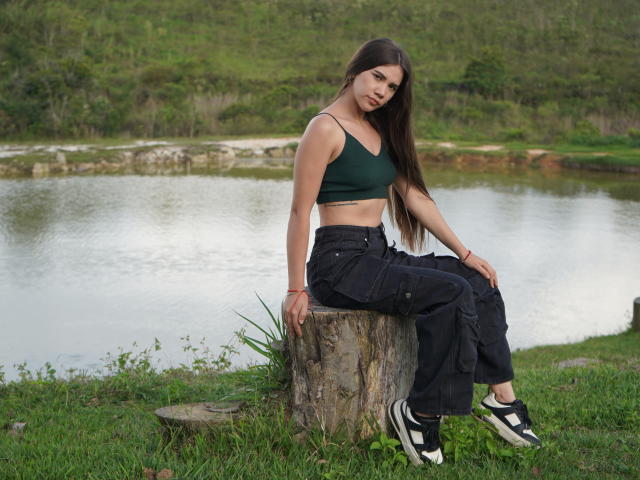 SashaHarding with long brown hair, green crop top, black pants, and black/white sneakers, sitting on a tree stump, looking at the camera, with a lake in the background.