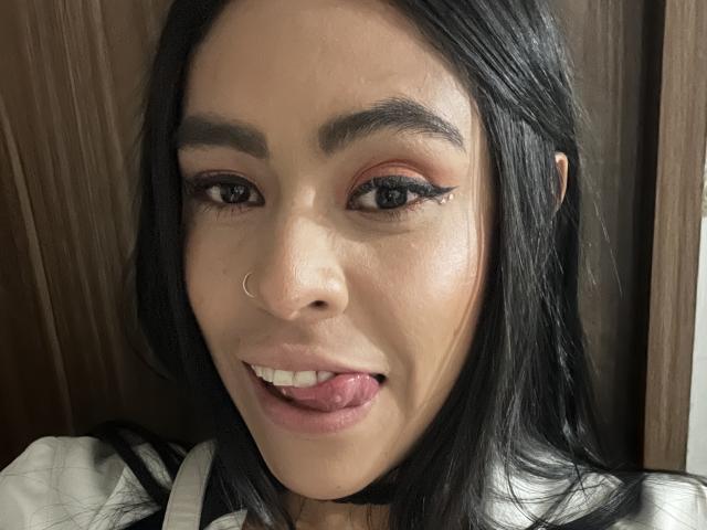 StellaBeckker, con cabello negro, piercing en la nariz y maquillaje, saca la lengua, mostrando su delineador de ojos, con las cejas levantadas. Su rostro está centrado, con un fondo de madera..