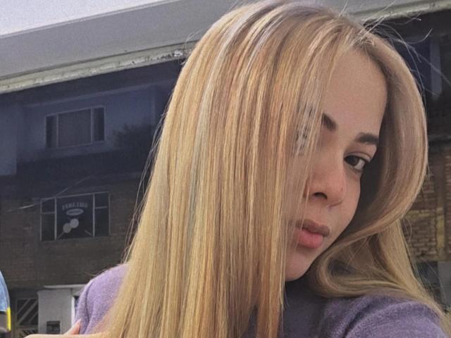 AlicePrice with long blonde hair, pouty lips, and subtle eye makeup, wearing a purple top, looking over her shoulder, her tits slightly visible under her shirt.
